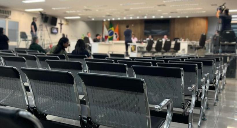 Sala de audiência com cadeiras vazias em primeiro plano e pessoas se preparando para uma reunião ao fundo.