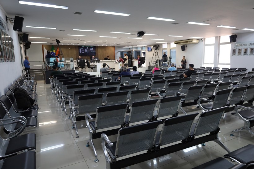 Sala de espera com cadeiras metálicas vazia, algumas pessoas sentadas e uma mesa à frente com pessoas em uma sessão ou reunião.