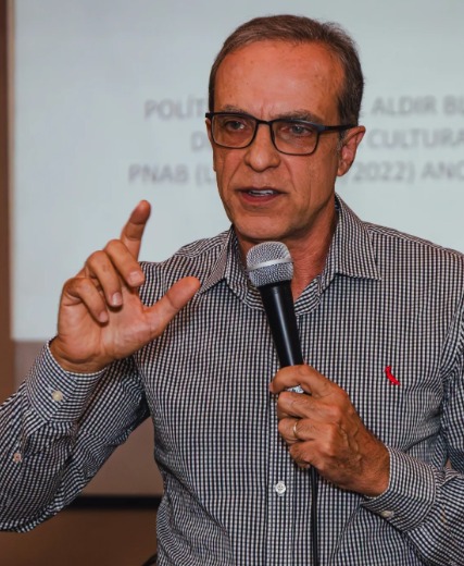 Homem de óculos segurando um microfone e gesticulando enquanto fala em um evento.