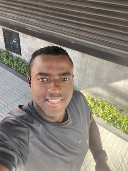 A man smiling and taking a selfie outdoors, wearing a black athletic shirt, with a landscaped area in the background.