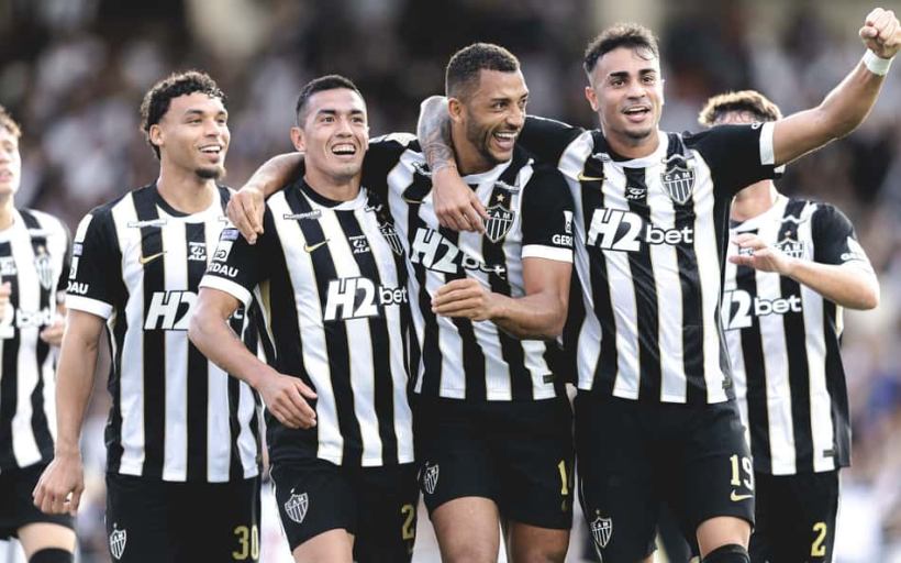 A group of four football players wearing black and white striped jerseys celebrate together on the field, smiling and showing enthusiasm.