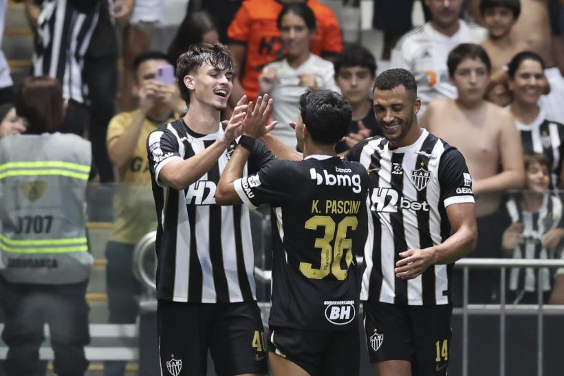 Three football players celebrating a goal on the field, wearing black and white striped jerseys, with enthusiastic fans in the background.
