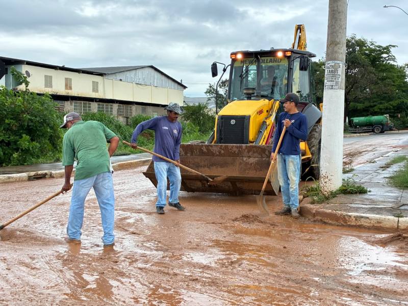 Três trabalhadores limpam uma rua enlameada com uma pá, enquanto uma máquina amarrotadora amarela está estacionada ao fundo.