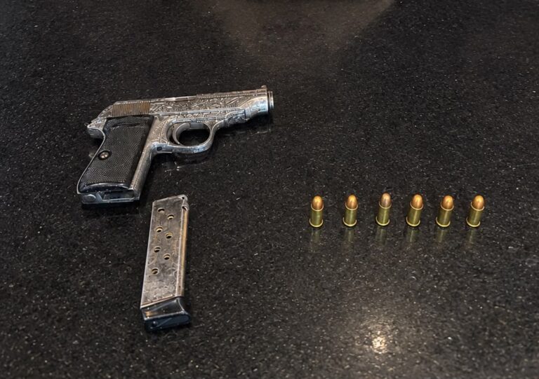 Uma pistola prateada com cabo de borracha ao lado de um carregador e sete balas de munição em uma superfície escura.