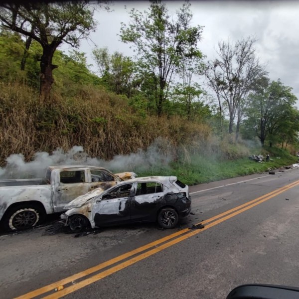 Veículos queimados ao lado da estrada, com fumaça saindo dos automóveis danificados.