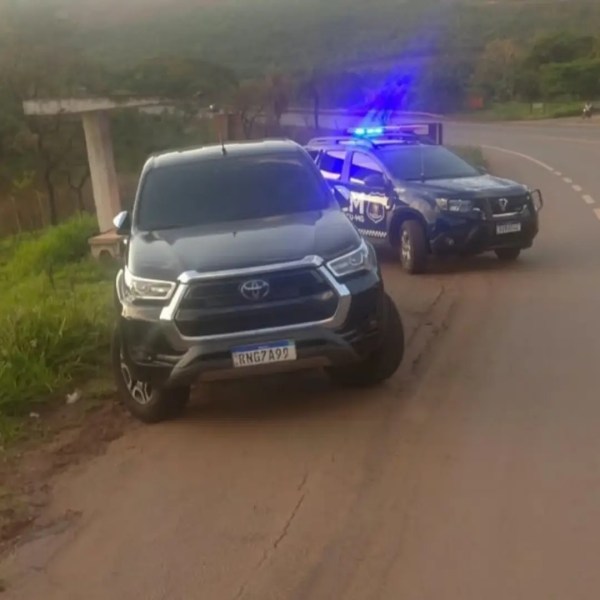 Uma caminhonete Toyota preta estacionada ao lado de uma viatura policial com luzes azuis ativadas em uma estrada.