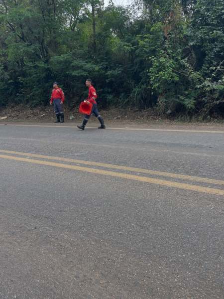 Duas pessoas vestindo roupas de combate a incêndio caminham pela beira da estrada, uma delas carregando um balde vermelho.