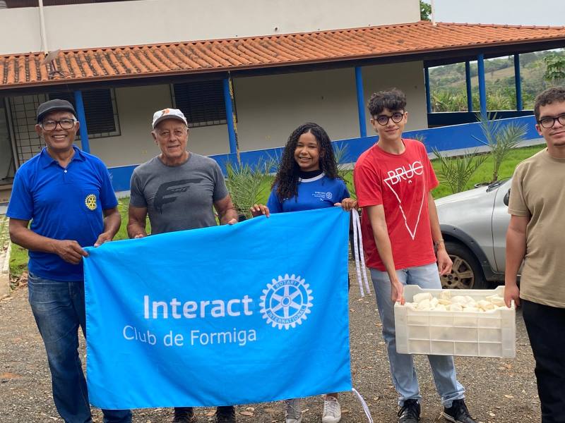 Grupo de pessoas sorrindo, segurando uma bandeira azul do Interact Club de Formiga e uma caixa com itens, em um ambiente externo com vegetação ao fundo.