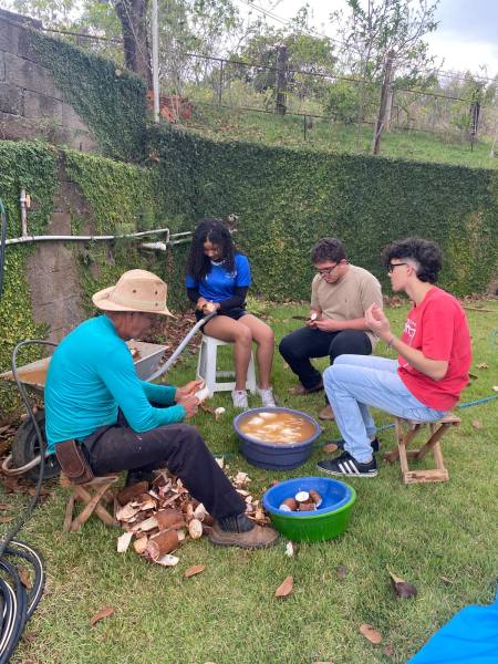 Um homem sentado, usando chapéu, descascando frutas ao ar livre, enquanto três jovens o observam e interagem. Ao fundo, uma parede coberta de vegetação e uma bacia com frutas em um ambiente natural.