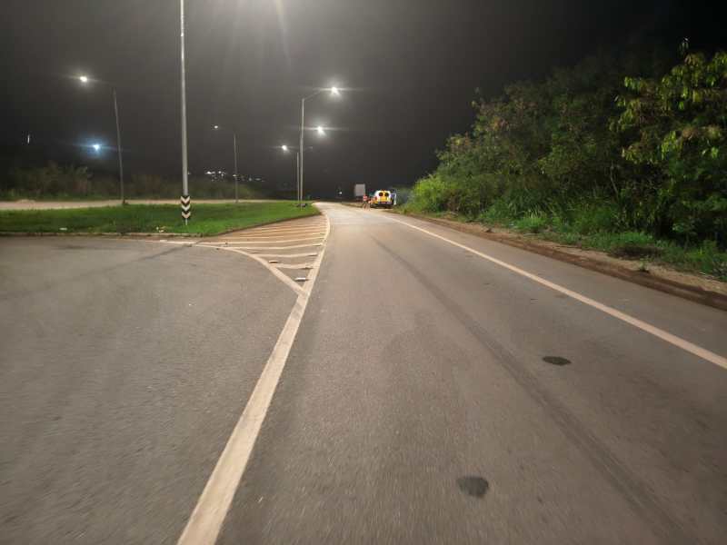 Imagem de uma estrada à noite com iluminação pública, mostrando uma curva e vegetação ao lado.