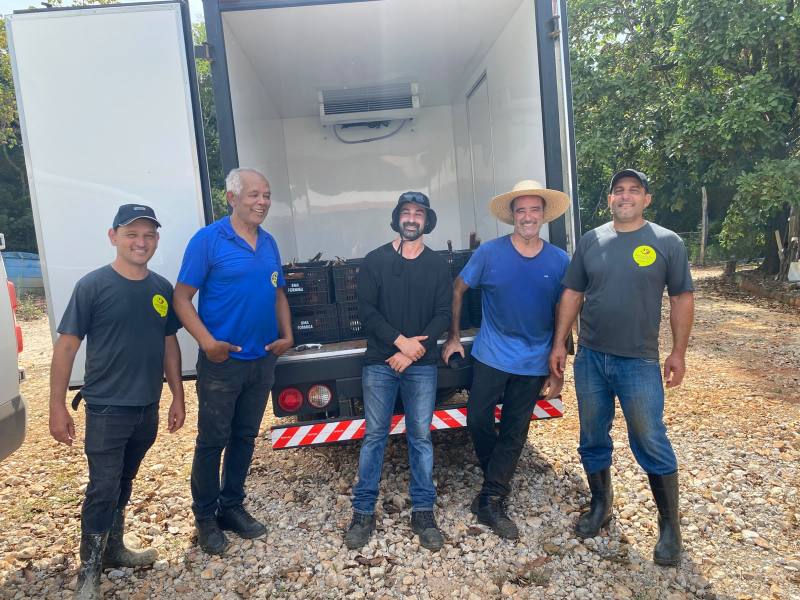 Cinco homens sorrindo em frente a um caminhão localizado em um ambiente rural, com caixas pretas visíveis dentro do veículo.