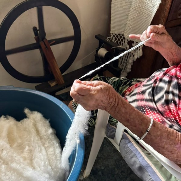 Uma pessoa idosa segurando fio enquanto trabalha com lã em uma roda de fiar, com um balde de lã ao lado.