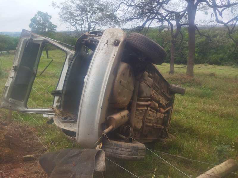 Carro capotado em um campo, com as rodas para cima e a porta aberta.