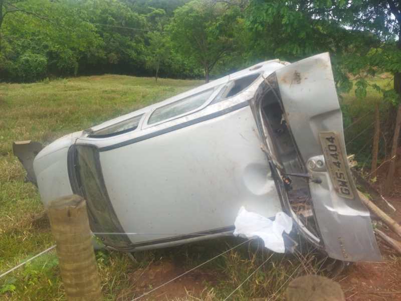 Carro capotado em um campo com árvores ao fundo.