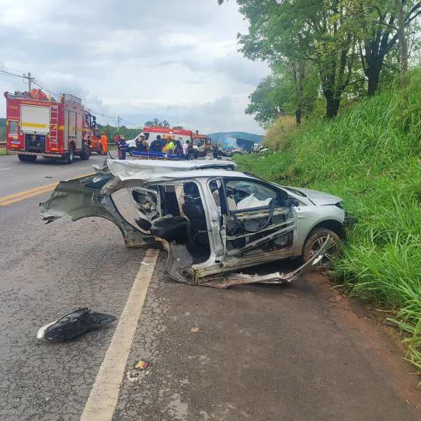 Veículo acidentado na beira da estrada, com danos significativos, incluindo a porta aberta. Equipes de emergência e viaturas ao fundo.