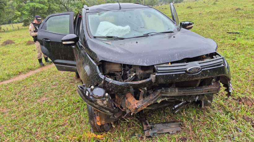 Carro preto envolvido em um acidente, com a frente danificada e a porta aberta, em um campo verde, enquanto um policial se aproxima.