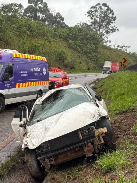 Carro acidentado na beira da estrada, com uma van de emergência e um caminhão ao fundo em um dia chuvoso.