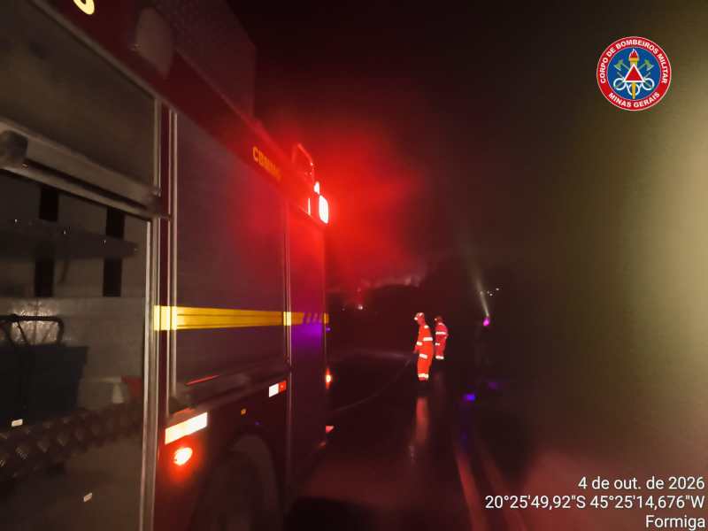 Imagem noturna de um caminhão de bombeiros com luzes vermelhas ativas, enquanto bombeiros em traje de segurança trabalham em uma estrada. Nevoeiro visível ao fundo.