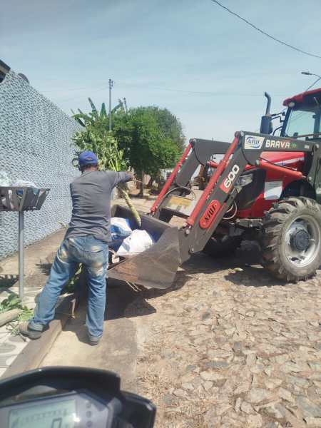 Um homem operando um trator com uma caçamba, carregando sacos e plantas em uma rua de paralelepípedos sob um céu claro.