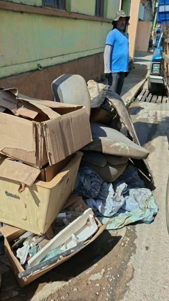 Montagem de lixo acumulado na rua, incluindo caixas de papelão e móveis, com um homem de boné ao fundo.