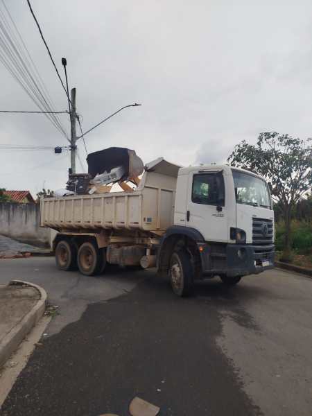 Caminhão de lixo branco, parcialmente carregado, na esquina de uma rua com árvores ao fundo.