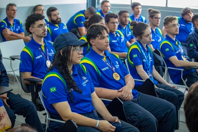 Grupo de jovens sentados em uma sala, todos usando camisetas azuis com detalhes em verde e amarelo, representando o Brasil.