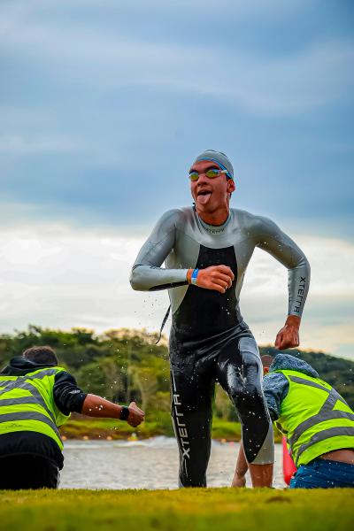 Atleta de triathlon saindo da água, utilizando traje de neoprene, exibindo uma expressão divertida com a língua para fora, enquanto recebe apoio de dois organizadores com coletes refletivos.