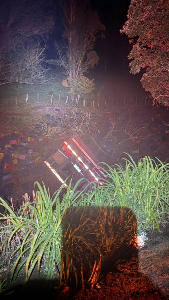 Caminhão tombado em um barranco à noite, rodeado por vegetação e caixas coloridas espalhadas ao redor.