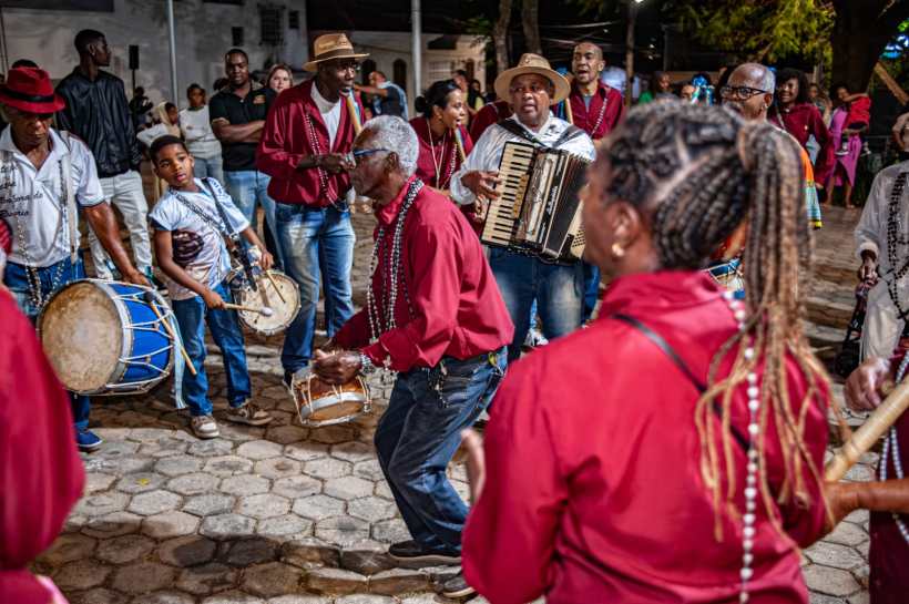 Grupo de músicos e dançarinos se apresentando em uma festa ao ar livre, com instrumentos como baterias, sanfona e flautas, todos vestindo roupas vermelhas e com colares de contas.