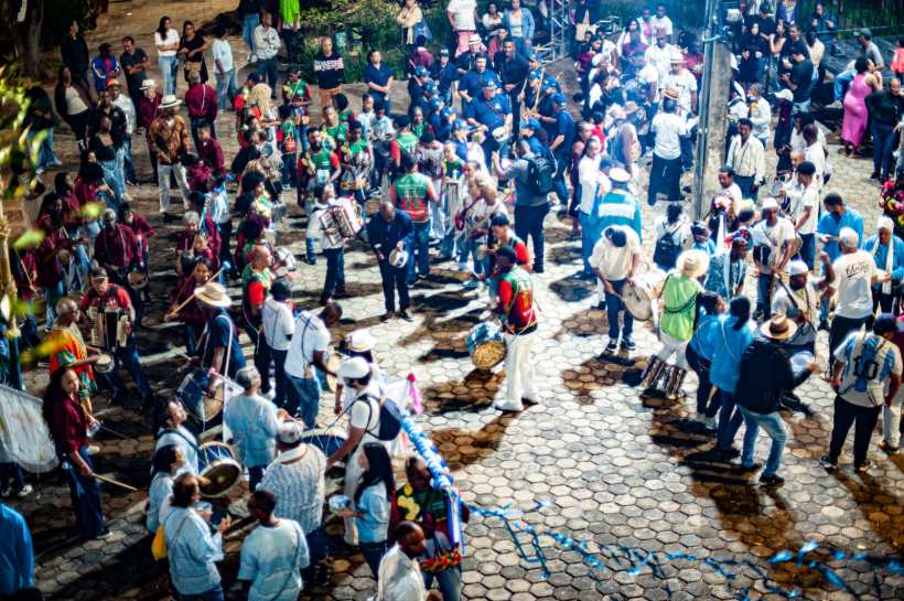 Uma multidão participa de uma celebração noturna em uma praça, com pessoas tocando instrumentos musicais e dançando em um ambiente festivo.