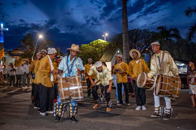 Grupo de pessoas dançando e tocando tambores em uma celebração noturna, com céu dramático ao fundo.