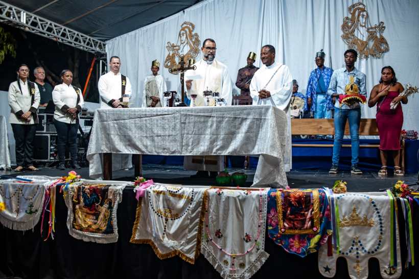 Celebration religiosa com pessoas em trajes formais ao redor de uma mesa, decorada com toalha branca, em um cenário festivo com várias bandeiras e ornamentos coloridos.