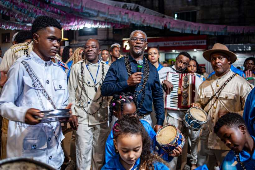 Grupo de pessoas se apresentando em um evento cultural, com foco em um homem falando ao microfone, enquanto outros tocam instrumentos musicais e crianças participam da celebração.