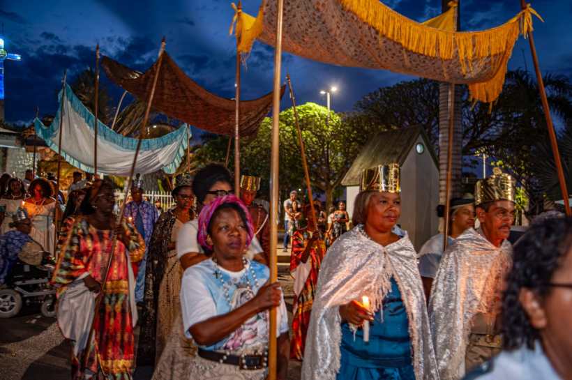 Multidão de pessoas em uma procissão, carregando bastões e pano, em um cenário noturno iluminado. Algumas pessoas usam vestimentas coloridas e chapéus dourados.
