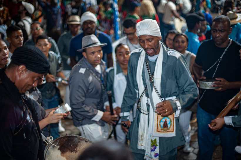 Grupo de pessoas em uma celebração cultural, com destaque para um homem trajando roupas tradicionais e cantando, rodeado por participantes que tocam instrumentos e assistem à cerimônia.