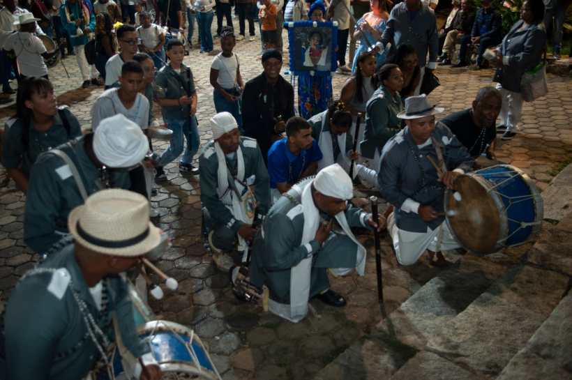 Grupo de músicos de pé e agachados tocando instrumentos em uma celebração cultural, com uma multidão assistindo ao fundo.