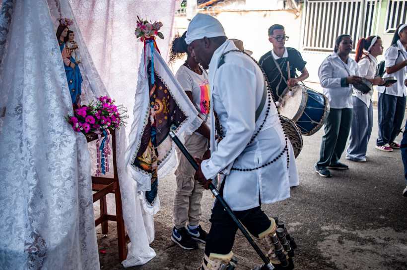 Homem vestido de branco carregando uma bandeira decorativa durante uma festividade, com imagens de santos ao fundo e grupo de pessoas dançando e tocando instrumentos.