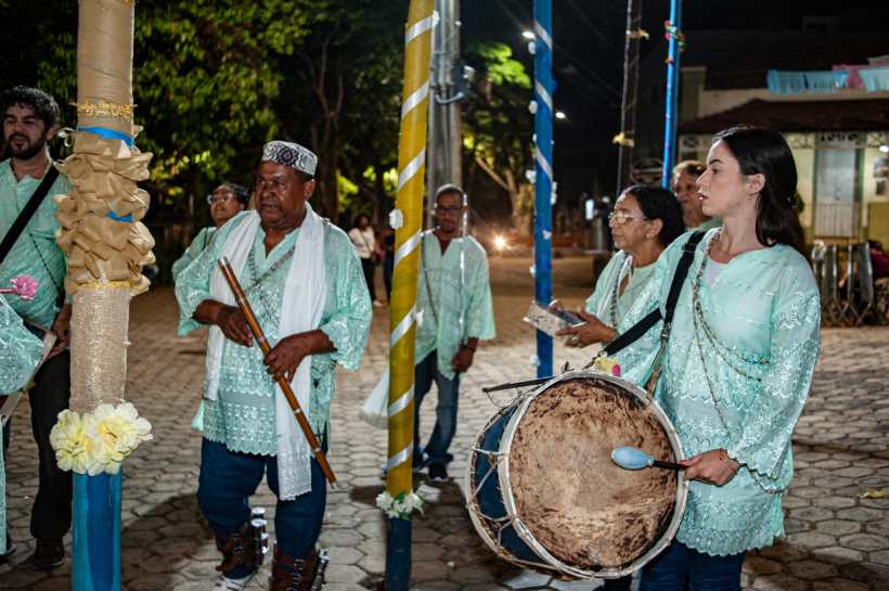 Grupo de pessoas vestindo roupas verdes, tocando instrumentos musicais e se reunindo em um ambiente noturno, cercados por postes decorados.