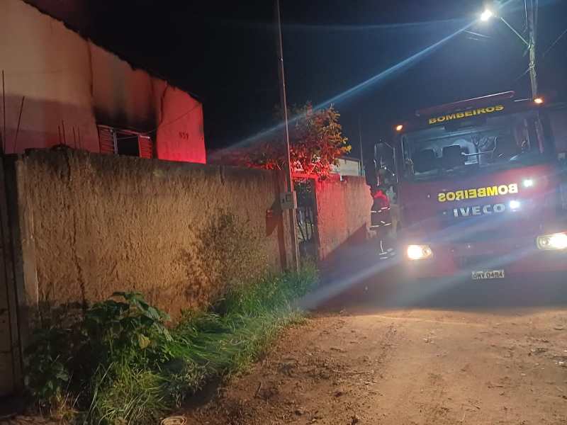Caminhão de bombeiros estacionado em uma rua à noite, com iluminação vermelha refletindo em uma parede e a casa ao fundo com sinais de incêndio.