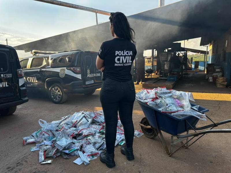 Agente da Polícia Civil de Minas Gerais observando uma pilha de sacos empilhados em um local com fumaça e veículos policiais ao fundo.