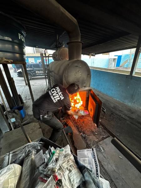 Um agente da polícia civil colocando documentos ou itens em um forno, com chamas visíveis na abertura do forno.
