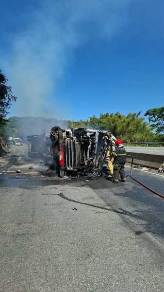 Caminhão tombado e em chamas na estrada, com bombeiros apagando o fogo ao fundo.