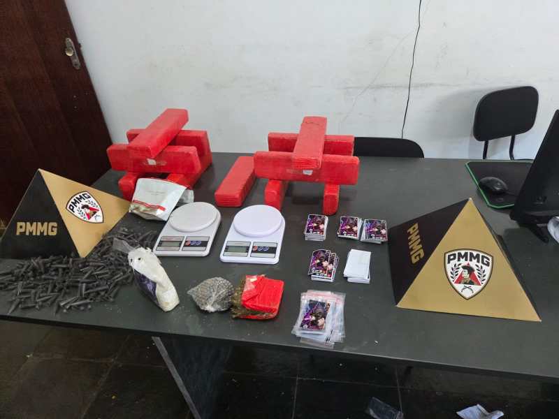 Mesa com blocos vermelhos, balanças digitais, sacos de substâncias, objetos diversos e placas com o emblema PMMG.