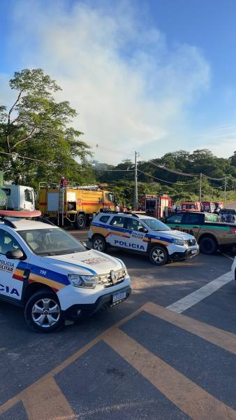 Vários veículos da polícia e do corpo de bombeiros em uma estrada, com fumaça ao fundo e árvores ao lado.