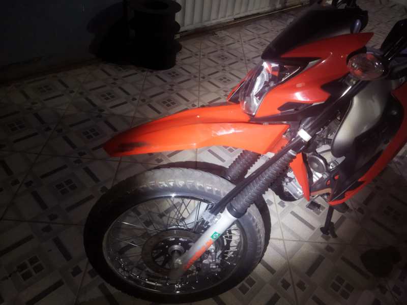 Moto de cor laranja, vista de perto, exibindo a roda dianteira e o guidão, em um ambiente interno com piso de azulejos.