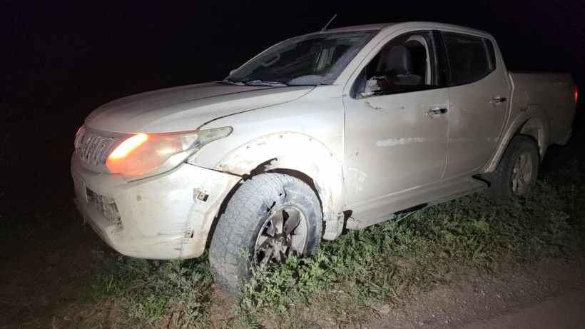 Pickup branca parada em um terreno com grama, à noite, com um dos pneus em uma posição inclinada.