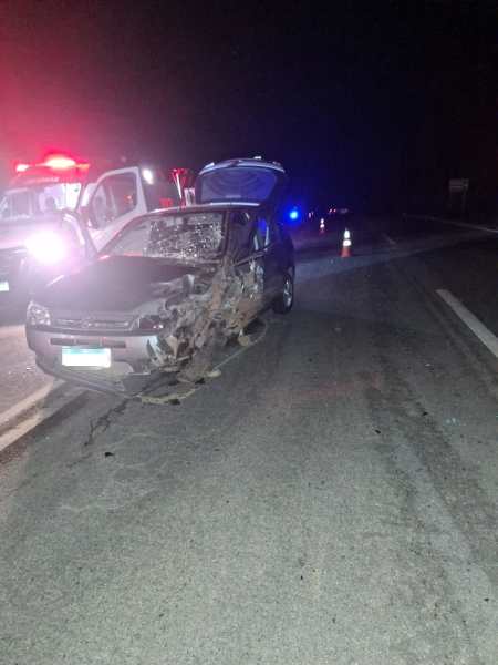 Carro danificado em uma estrada à noite, com sinais de acidente e veículos de emergência ao fundo.
