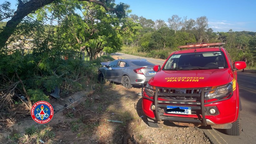 Veículo dos bombeiros parados ao lado de um carro acidentado em uma estrada, com vegetação ao redor.