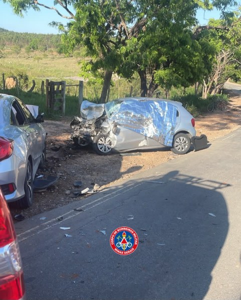A imagem mostra um acidente de carro com um veículo danificado com a parte frontal colidida e coberta com uma lona prateada, estacionado na beira da estrada. Outro carro está visível ao lado, também danificado, em um ambiente rural com árvores ao fundo.
