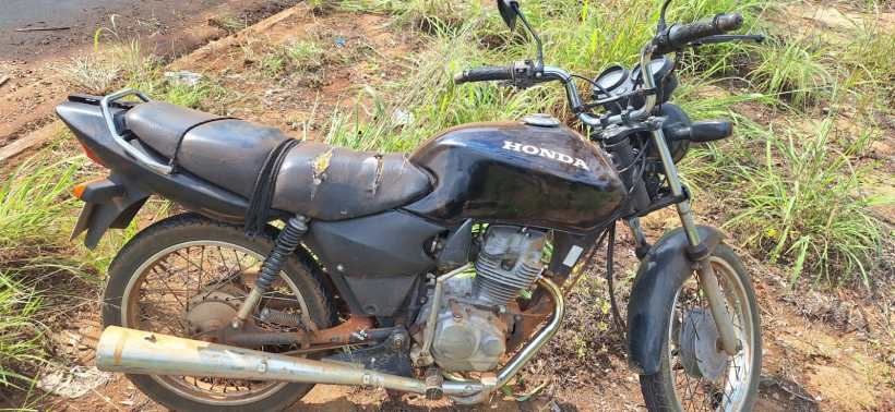 Uma moto Honda preta, velha e desgastada, estacionada em um terreno coberto de grama.